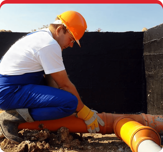 A man in an orange helmet kneels down next to pipes.